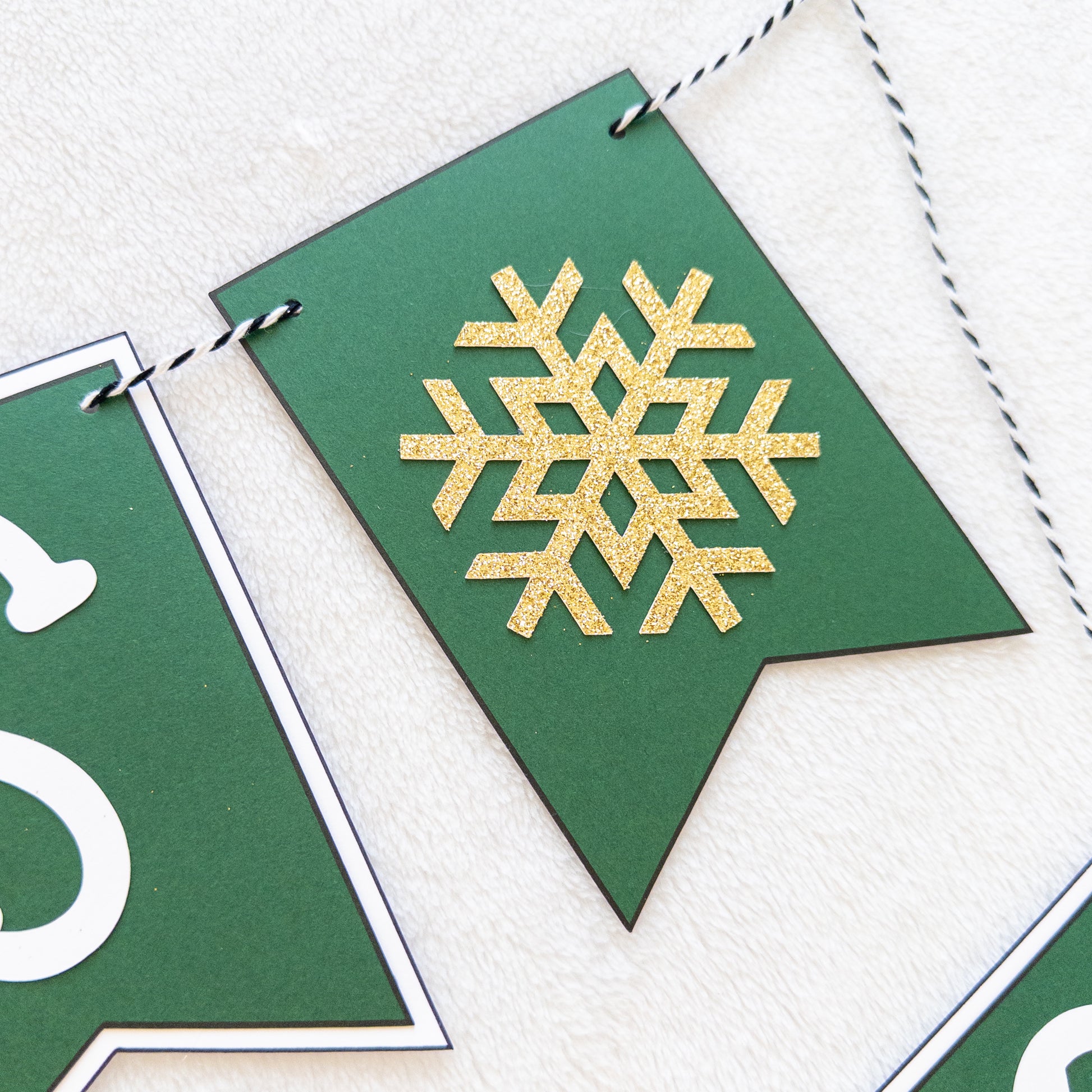 A banner with the text 'BABY IT'S COLD OUTSIDE' in capital letters, hanging by a string. The letters are white on green and white bunting, and there are decorative snowflake-like patterns at the ends of the strings.