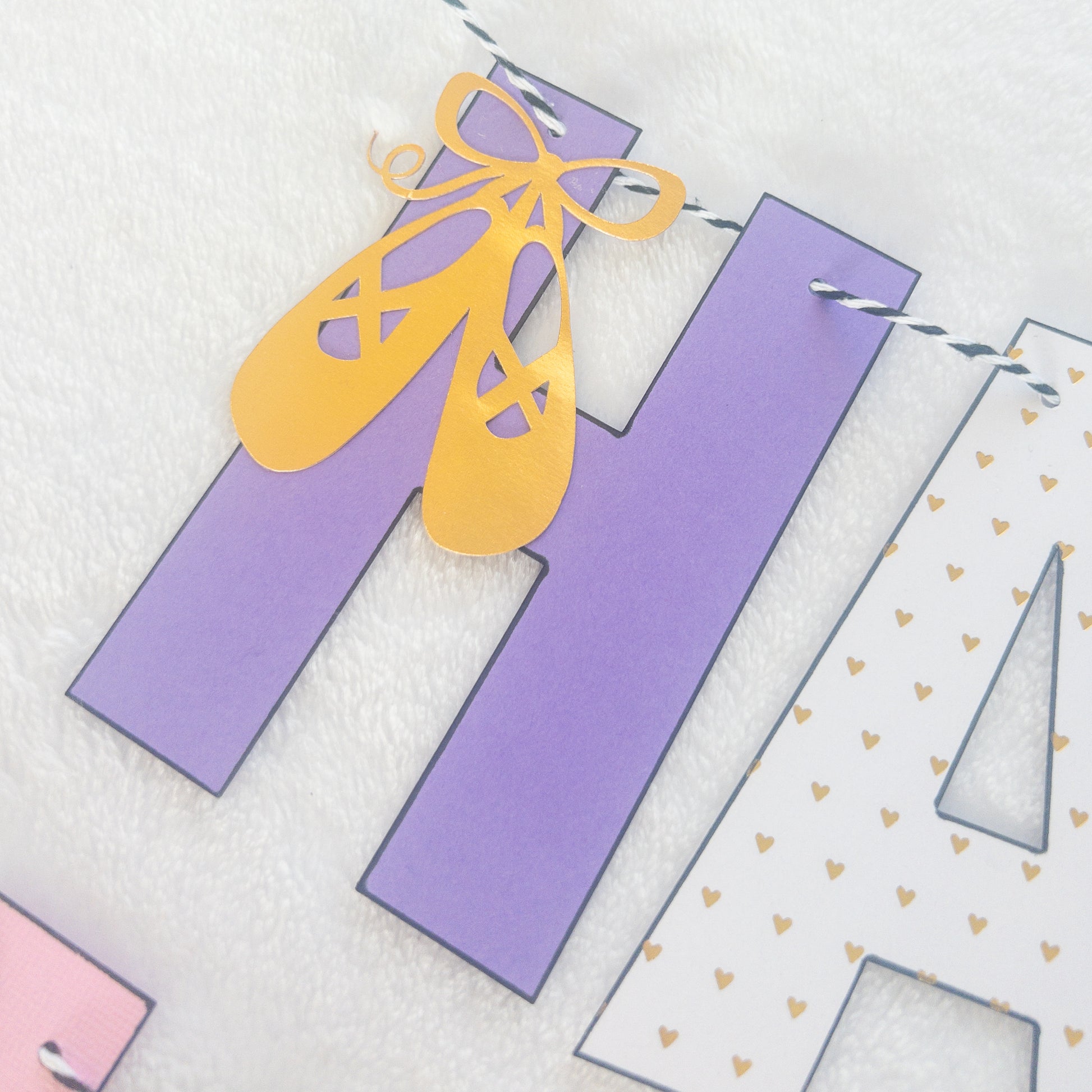 A Happy Birthday banner with the words 'HAPPY BIRTHDAY' in bold letters, decorated with small purple, pink, and yellow details, such as ballet shoes and a tutu, strung on a string.