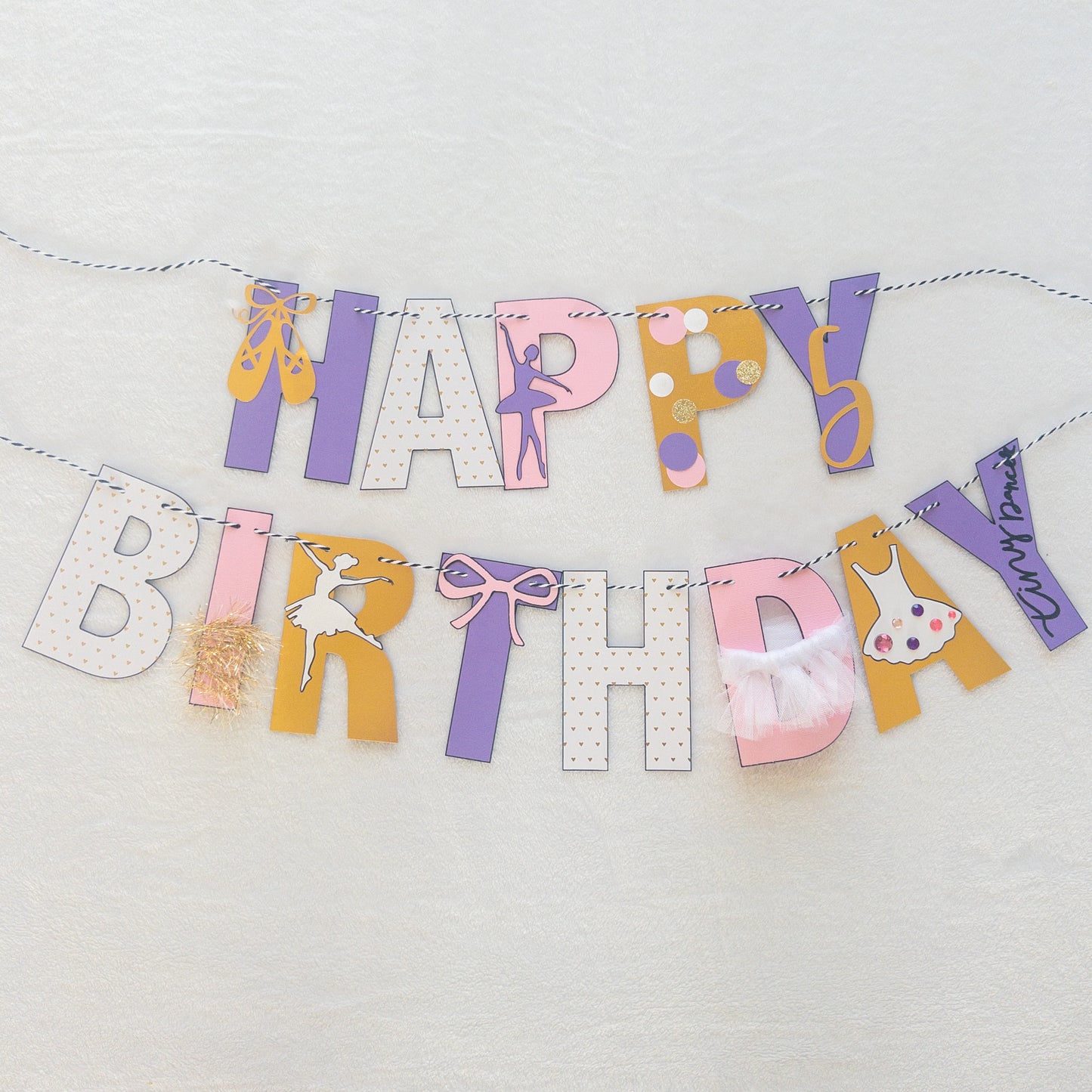 A Happy Birthday banner with the words 'HAPPY BIRTHDAY' in bold letters, decorated with small purple, pink, and yellow details, such as ballet shoes and a tutu, strung on a string.