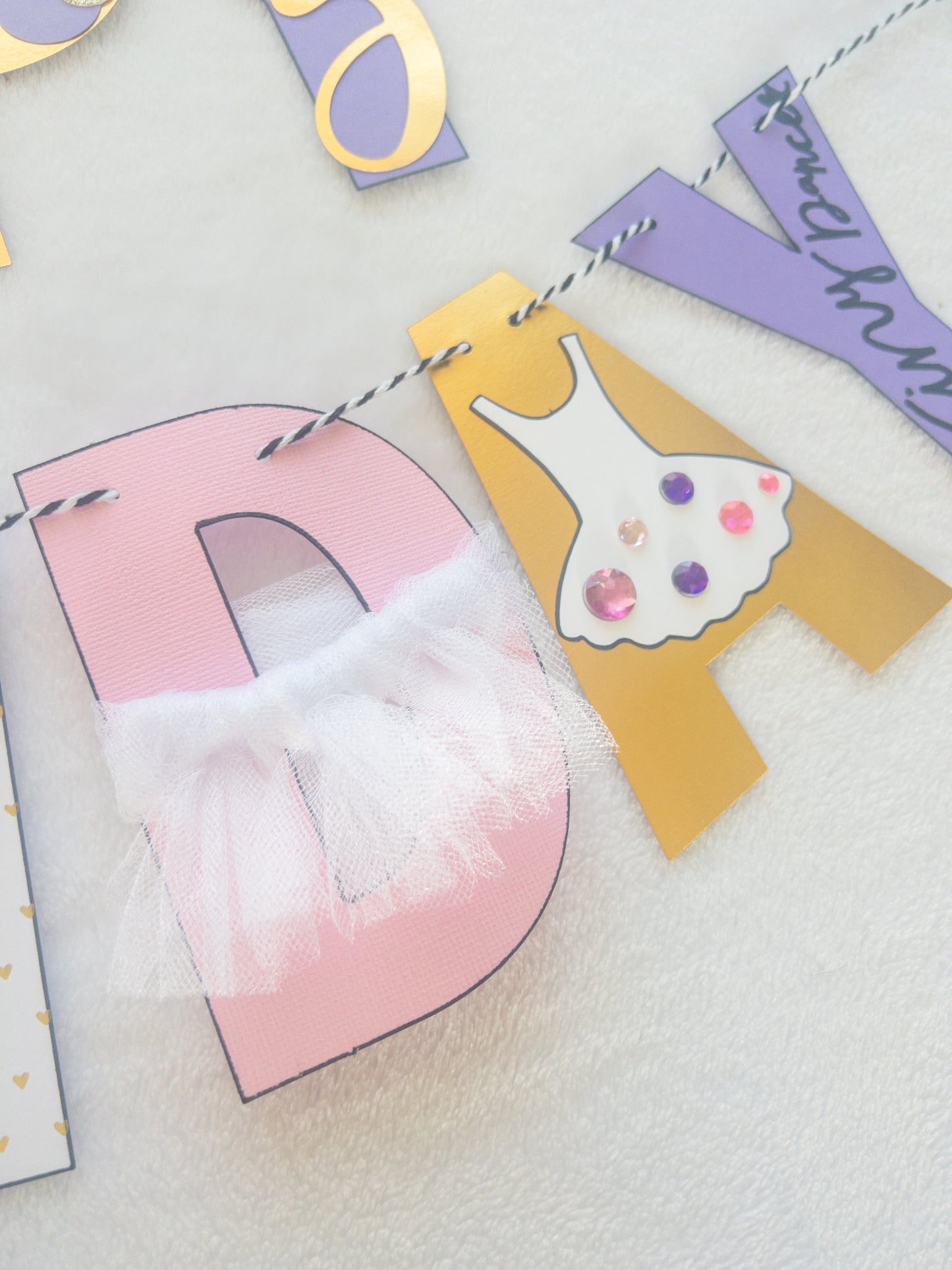 A Happy Birthday banner with the words 'HAPPY BIRTHDAY' in bold letters, decorated with small purple, pink, and yellow details, such as ballet shoes and a tutu, strung on a string.