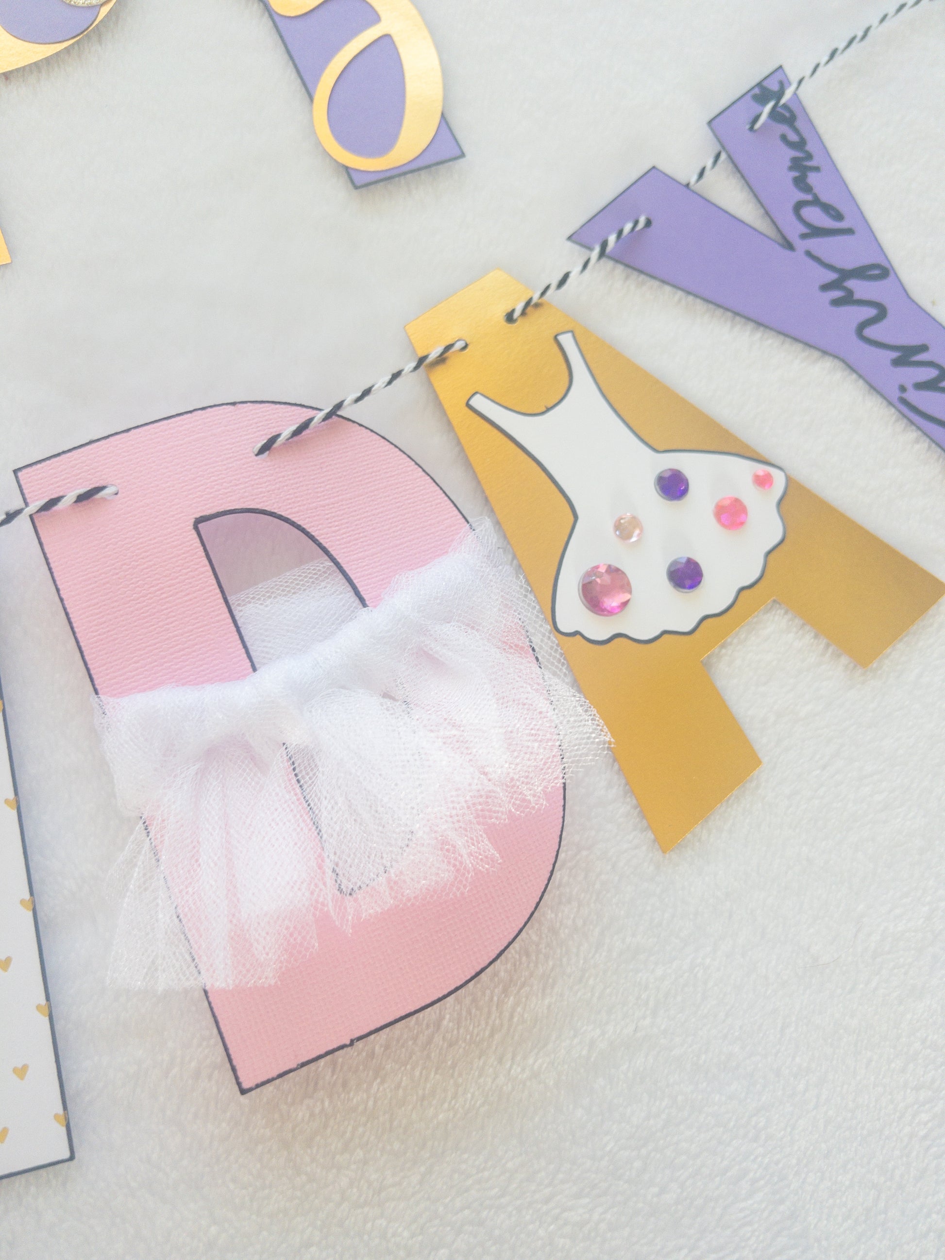 A Happy Birthday banner with the words 'HAPPY BIRTHDAY' in bold letters, decorated with small purple, pink, and yellow details, such as ballet shoes and a tutu, strung on a string.