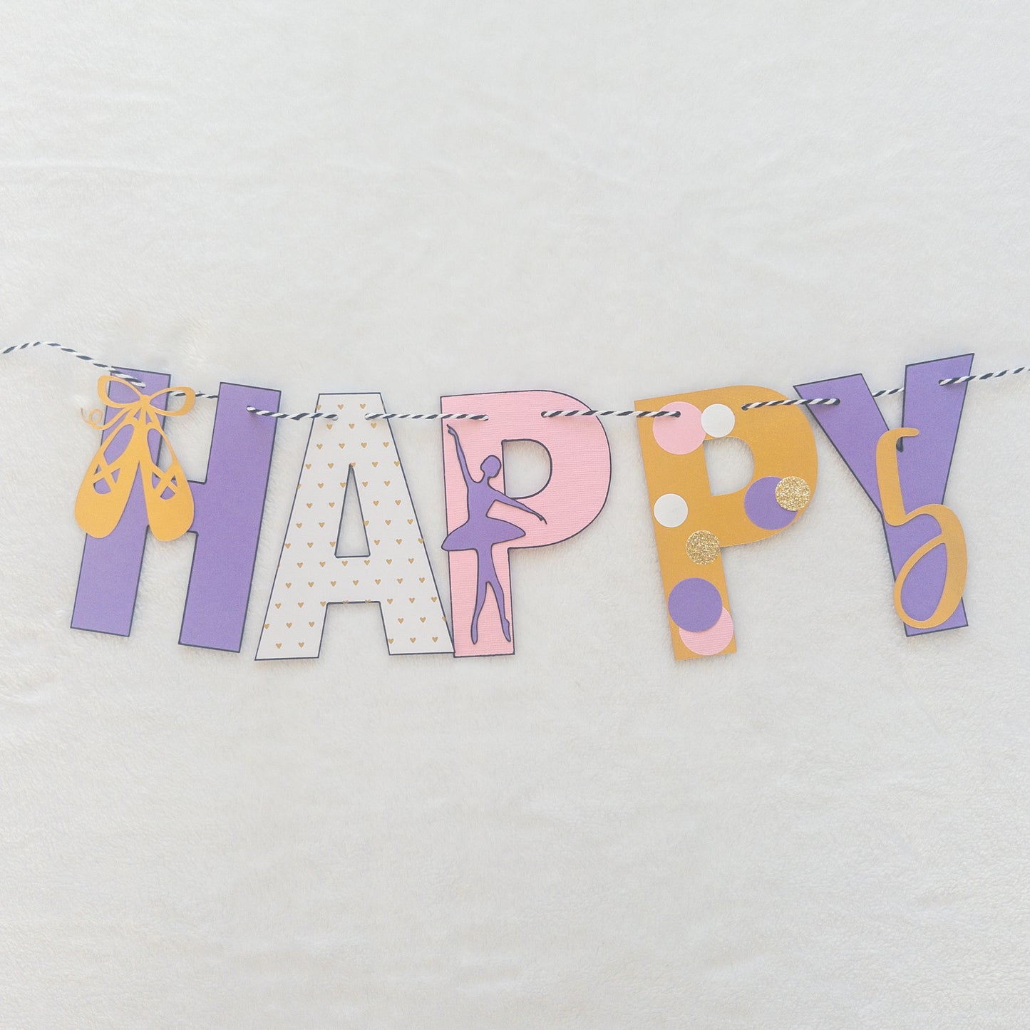 A Happy Birthday banner with the words 'HAPPY BIRTHDAY' in bold letters, decorated with small purple, pink, and yellow details, such as ballet shoes and a tutu, strung on a string.