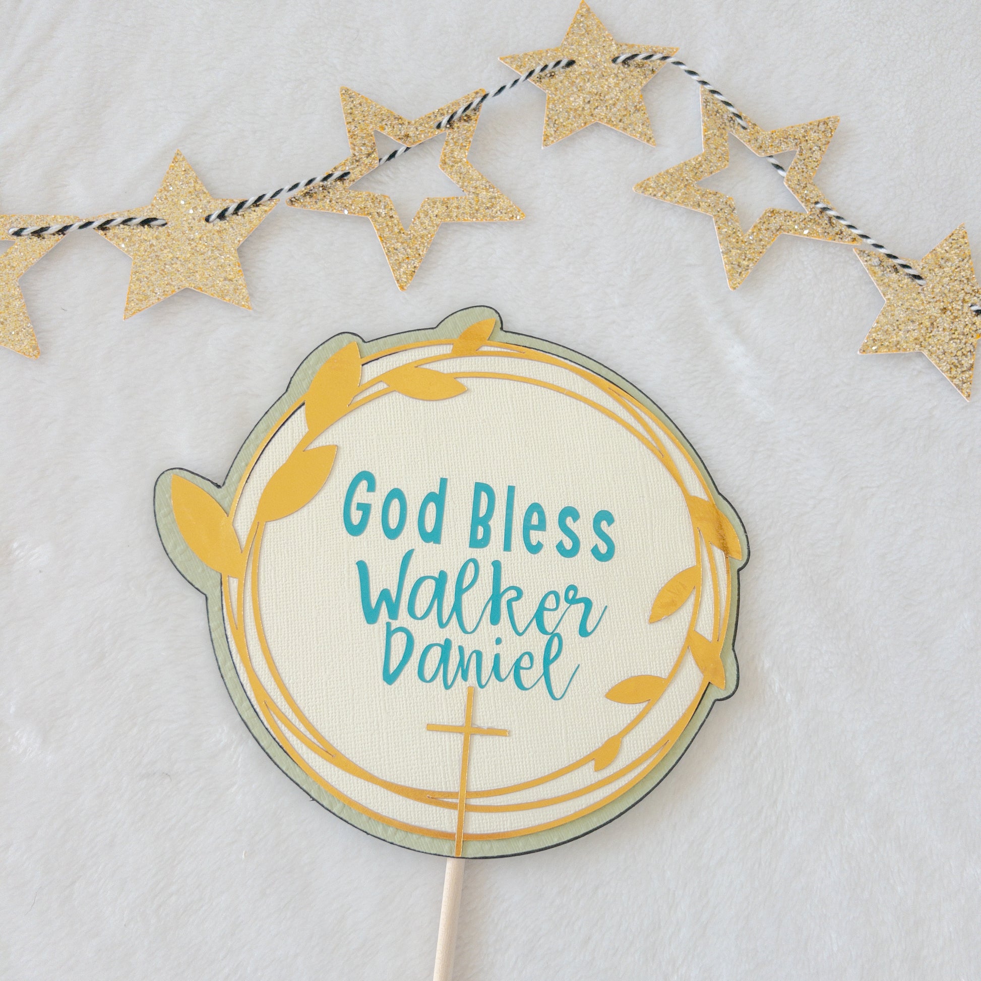 A cake topper with a gold background, teal text, and decorative stars, designed for a baptism or christening celebration.