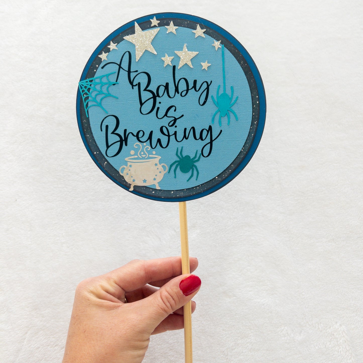 A circular cake topper with a blue background, featuring the text 'A BABY IS BREWING' in a cursive font, Halloween-themed decorations such as a spider, spider web, and a cauldron , and stars.