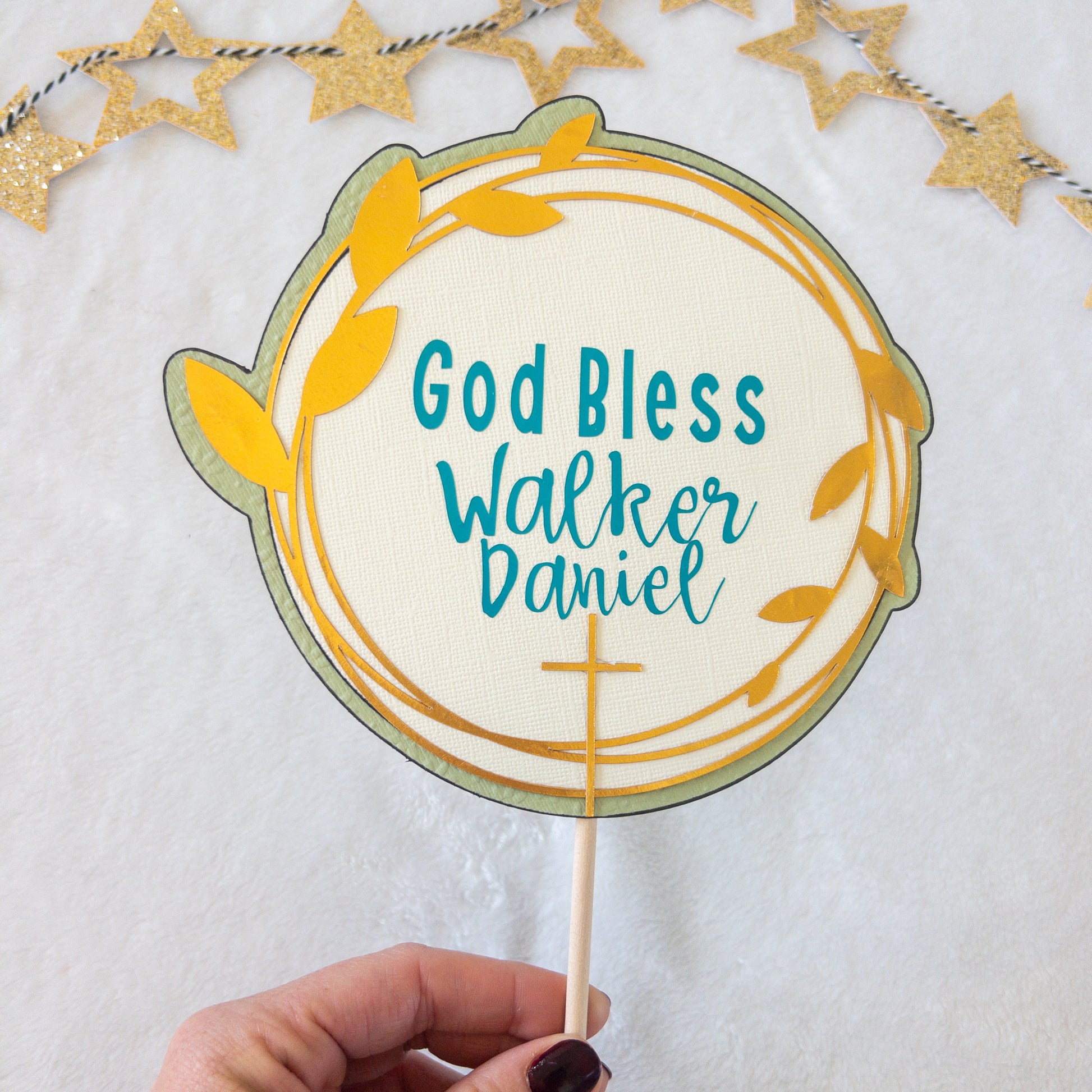 A cake topper with a gold background, teal text, and decorative stars, designed for a baptism or christening celebration.