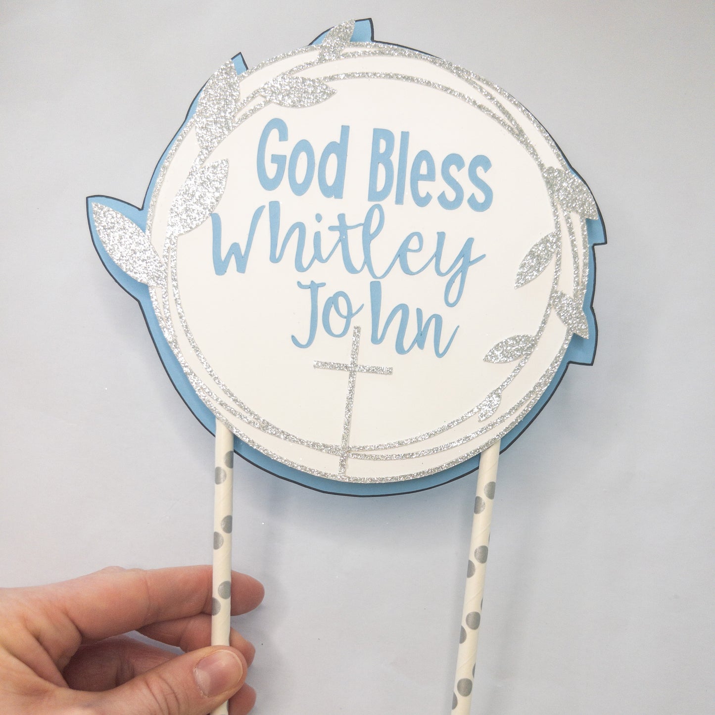 A cake topper with a white background, blue text, and decorative flowers, designed for a baptism or christening celebration.