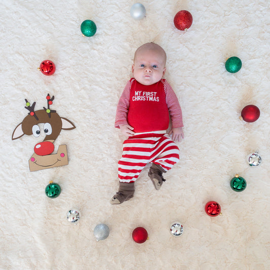 A baby lying on a white furry surface with a Christmas-themed backdrop, including decorative red and green balls and a cartoon reindeer.
