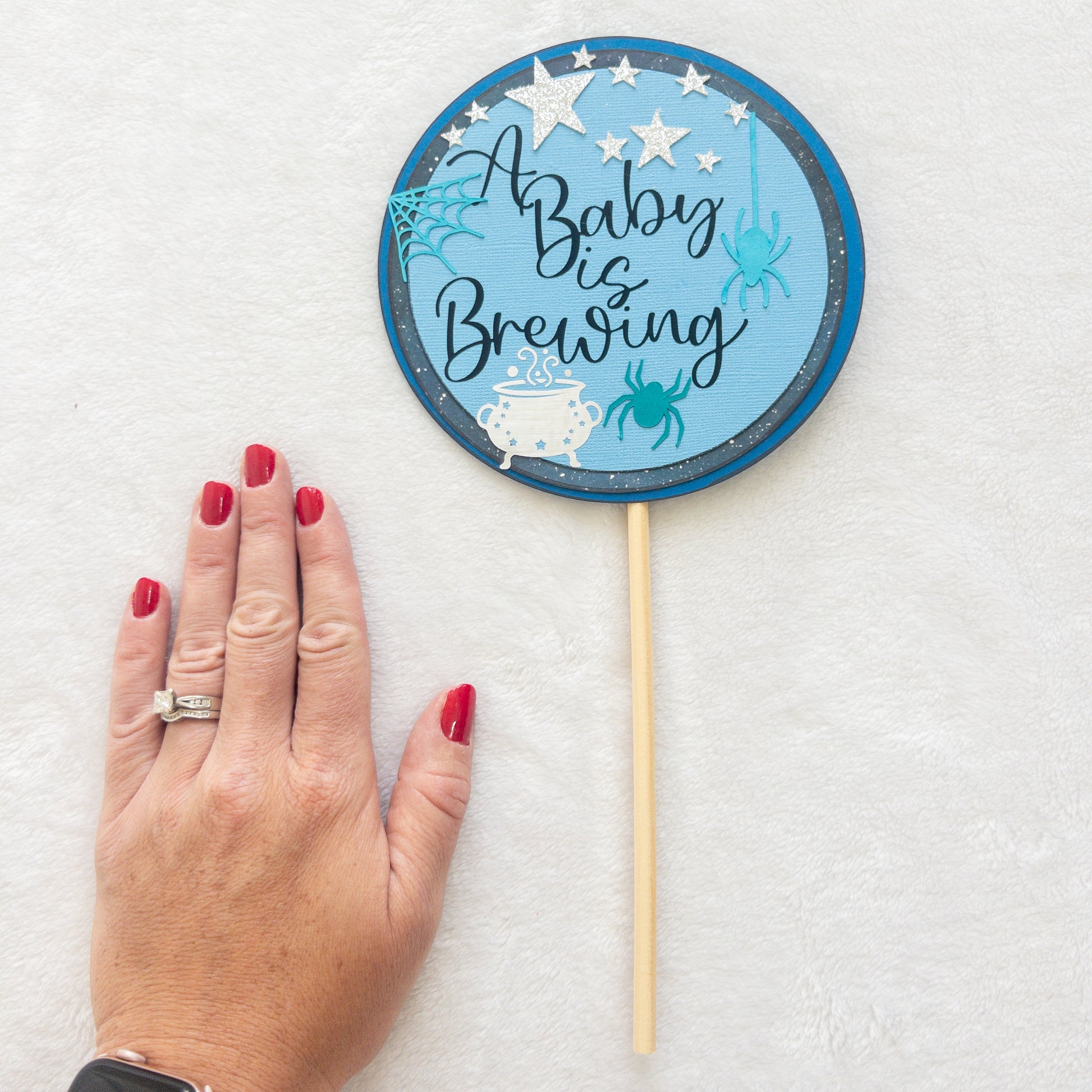A circular cake topper with a blue background, featuring the text 'A BABY IS BREWING' in a cursive font, Halloween-themed decorations such as a spider, spider web, and a cauldron , and stars.