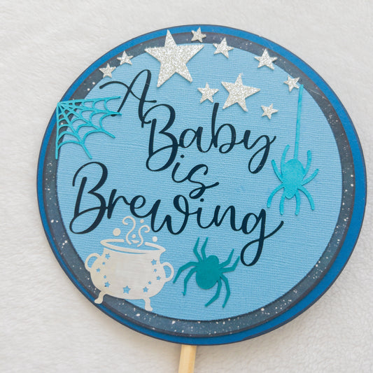 A circular cake topper with a blue background, featuring the text 'A BABY IS BREWING' in a cursive font, Halloween-themed decorations such as a spider, spider web, and a cauldron , and stars.