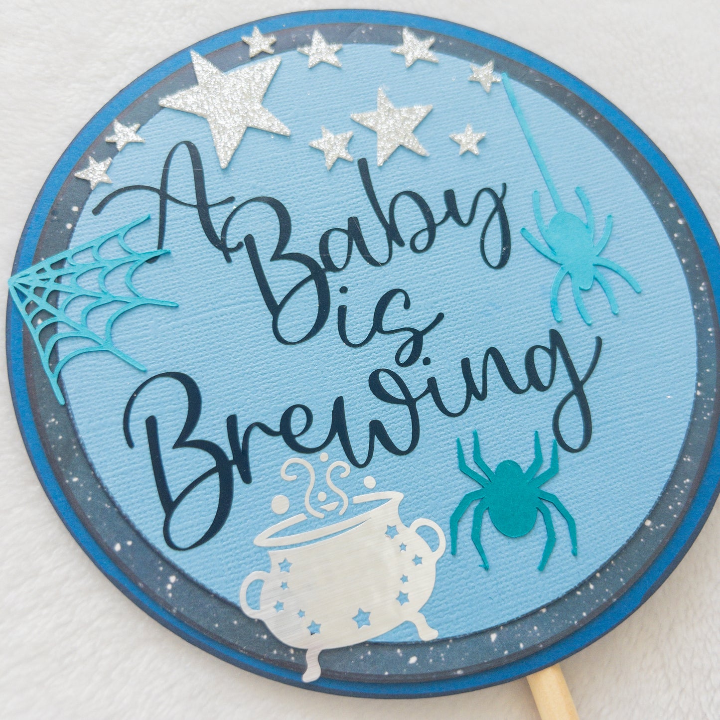 A circular cake topper with a blue background, featuring the text 'A BABY IS BREWING' in a cursive font, Halloween-themed decorations such as a spider, spider web, and a cauldron , and stars.