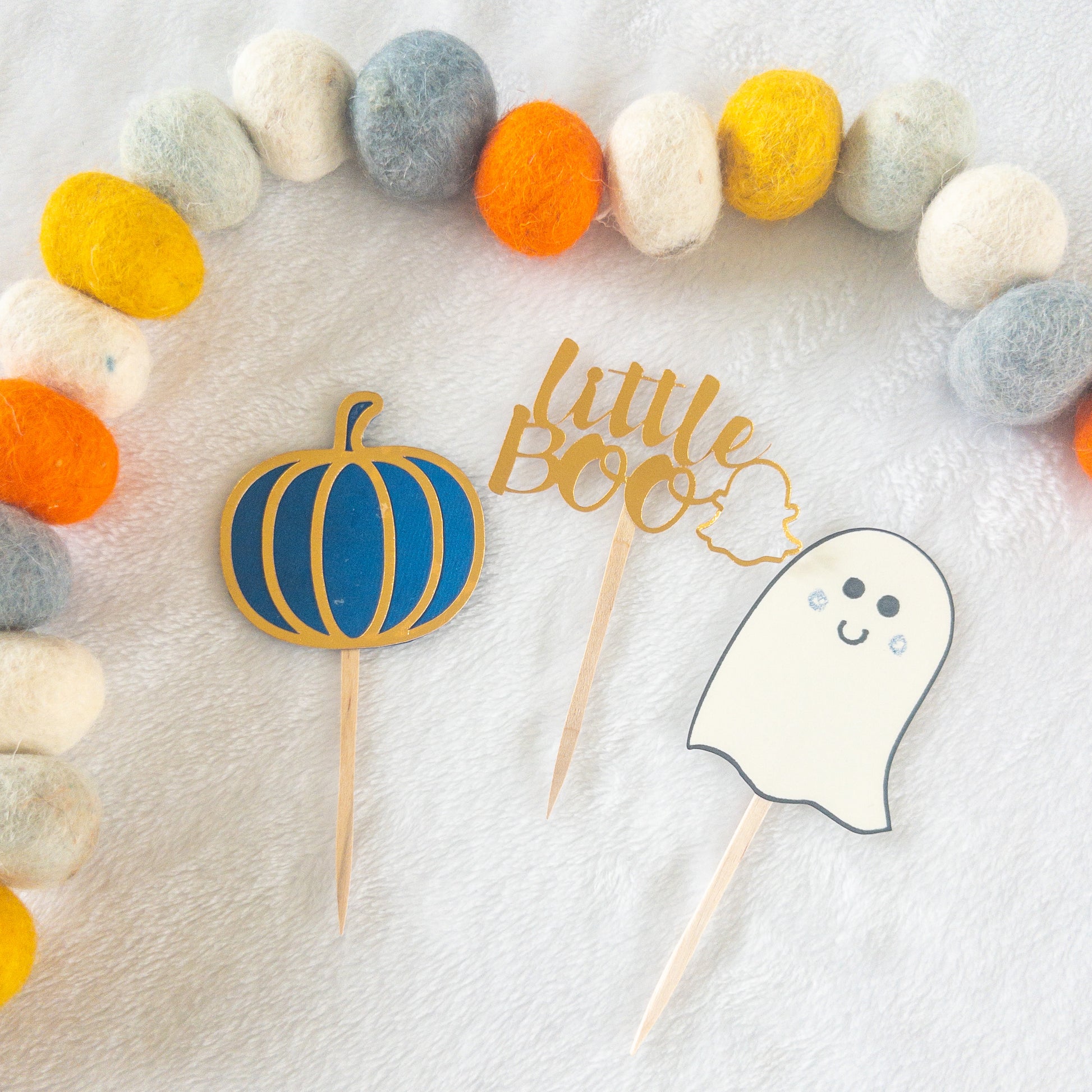 Cupcake toppers with wooden sticks, one designed as a ghost, 'Little Boo' text, and the other shaped like a pumpkin