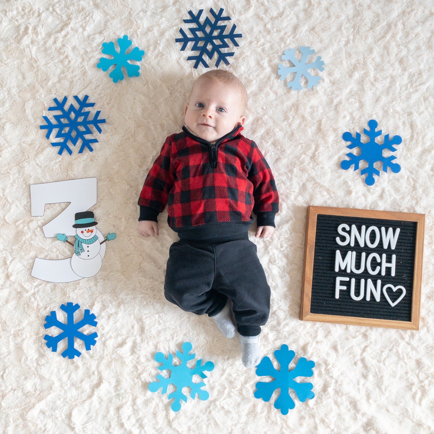 A baby laying on a furry white blanket next to snowflakes and a sign that says ‘snow much fun’