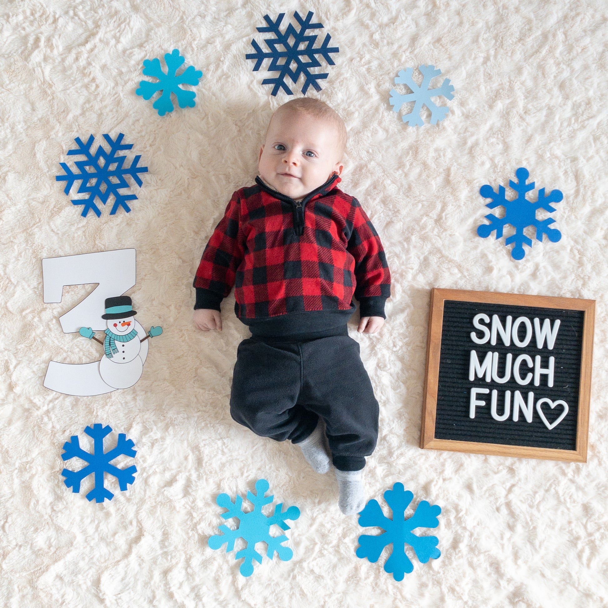 A baby laying on a furry white blanket next to snowflakes and a sign that says ‘snow much fun’