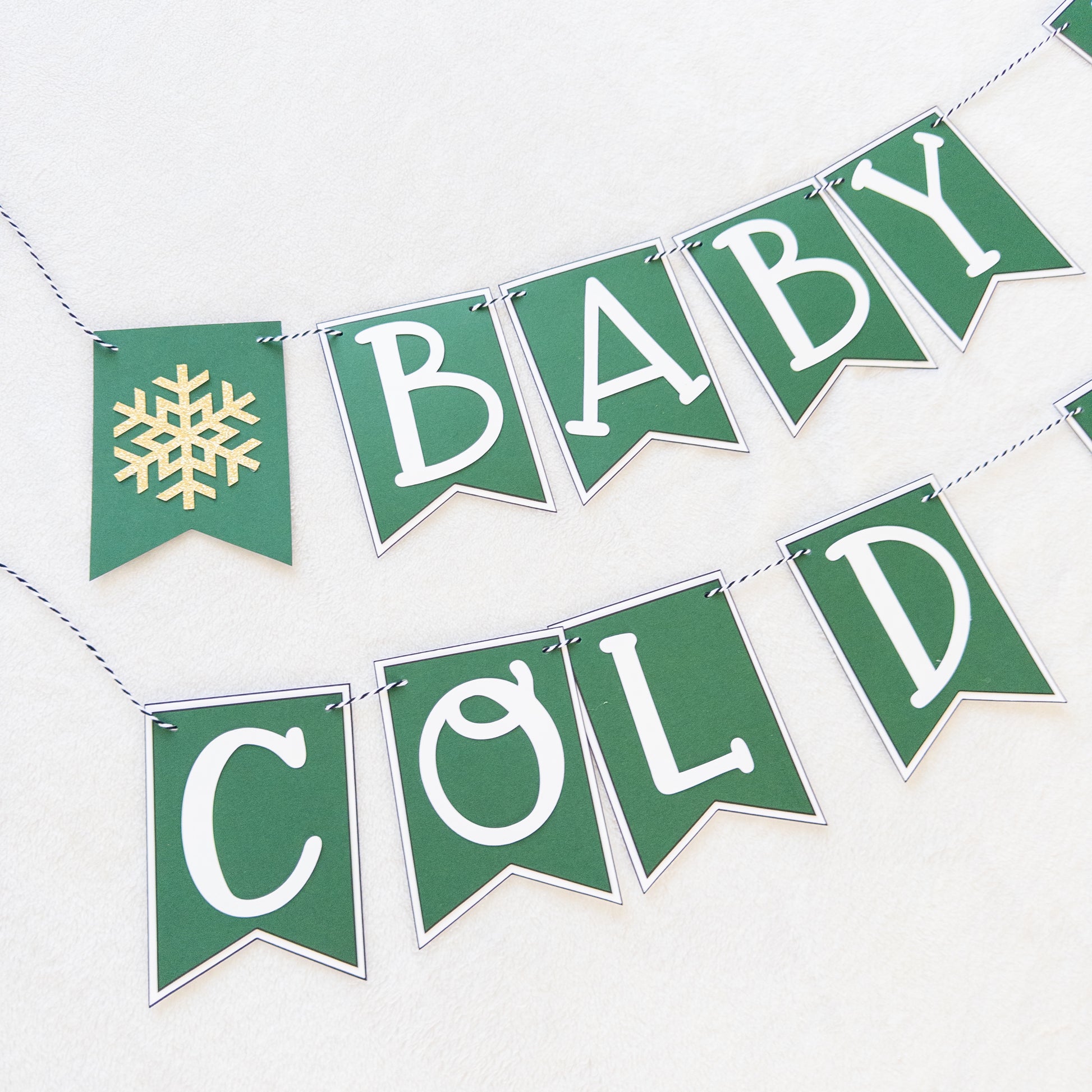 A banner with the text 'BABY IT'S COLD OUTSIDE' in capital letters, hanging by a string. The letters are white on green and white bunting, and there are decorative snowflake-like patterns at the ends of the strings.