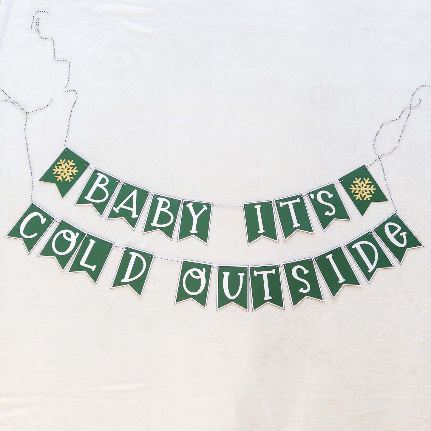A banner with the text 'BABY IT'S COLD OUTSIDE' in capital letters, hanging by a string. The letters are white on green and white bunting, and there are decorative snowflake-like patterns at the ends of the strings.