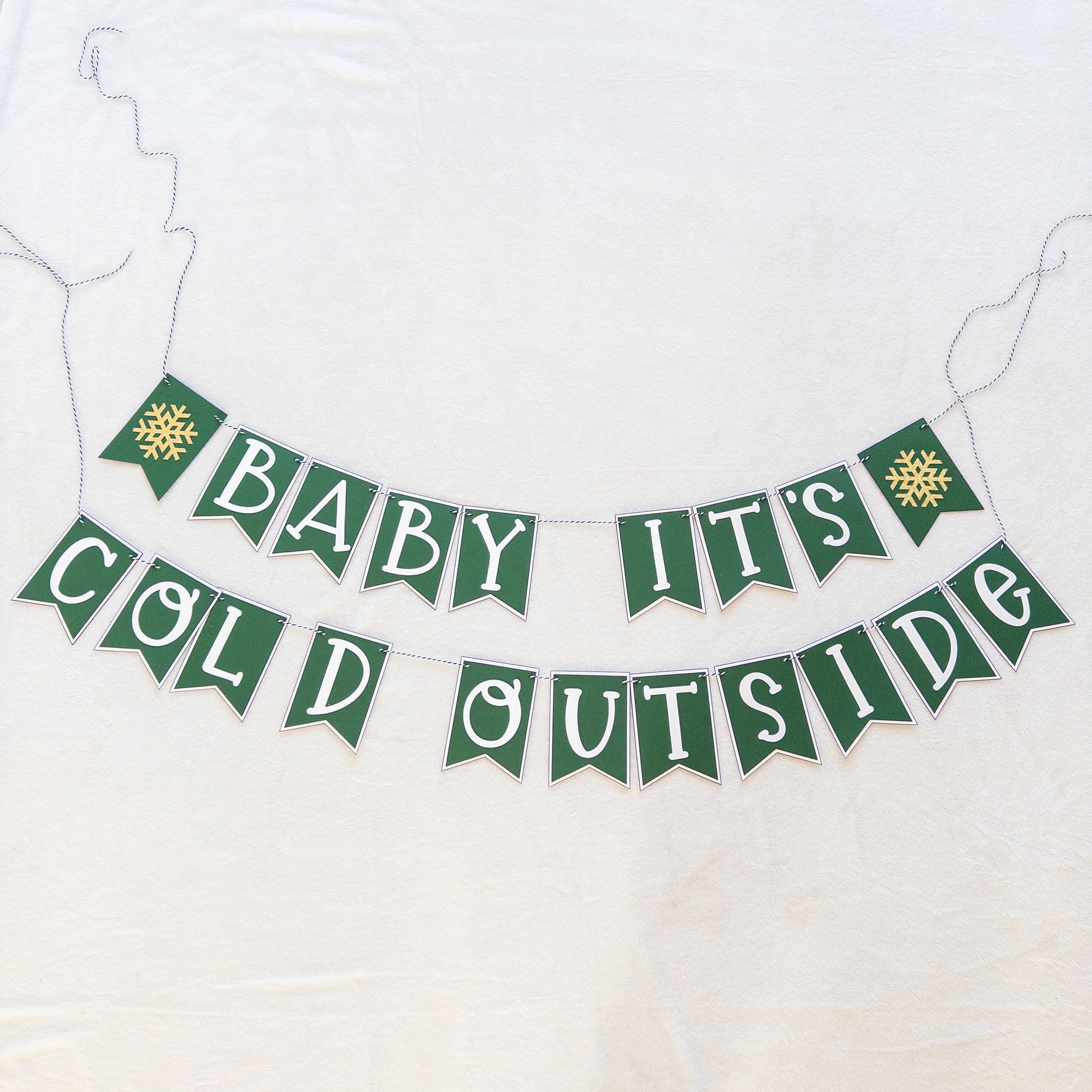 A banner with the text 'BABY IT'S COLD OUTSIDE' in capital letters, hanging by a string. The letters are white on green and white bunting, and there are decorative snowflake-like patterns at the ends of the strings.