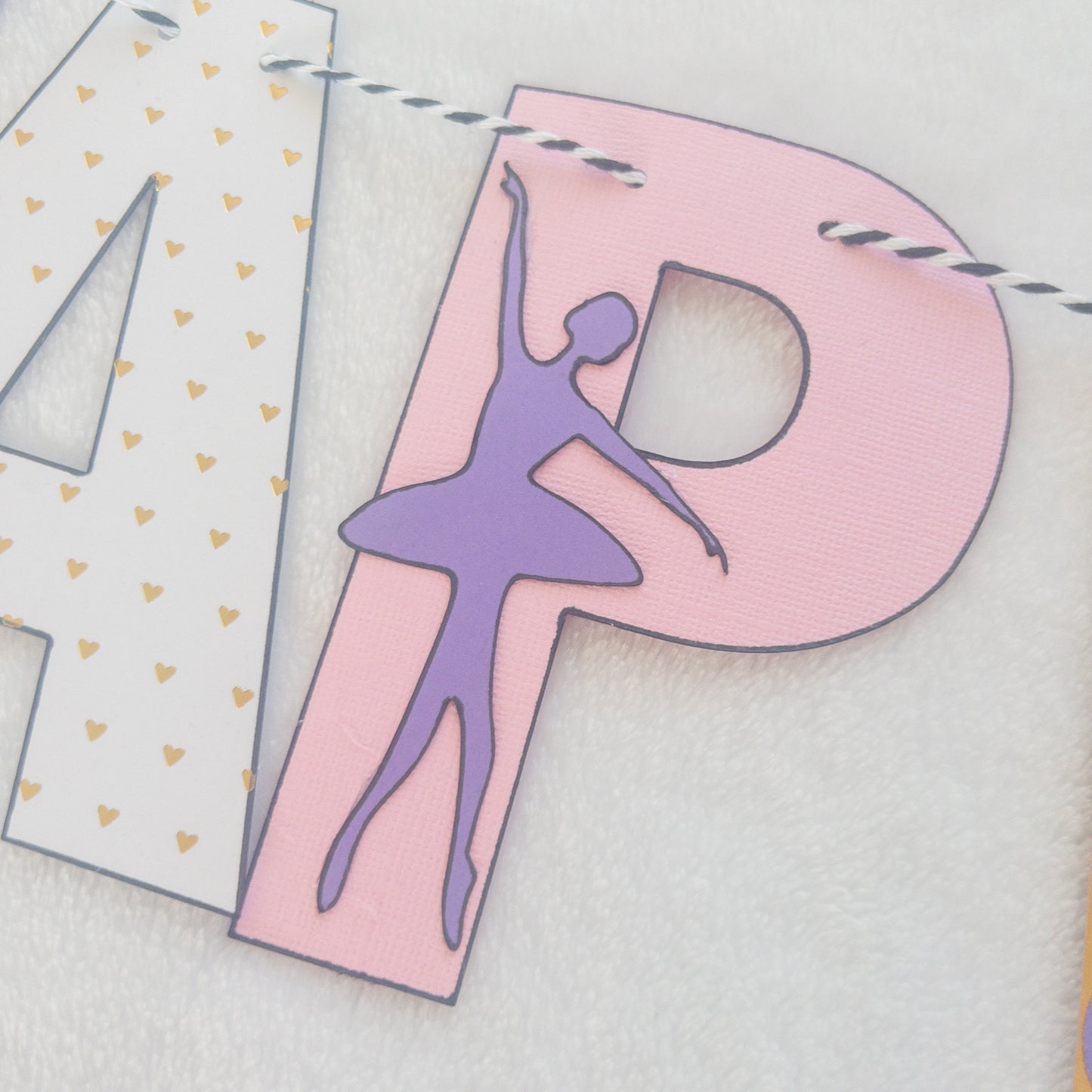 A Happy Birthday banner with the words 'HAPPY BIRTHDAY' in bold letters, decorated with small purple, pink, and yellow details, such as ballet shoes and a tutu, strung on a string.